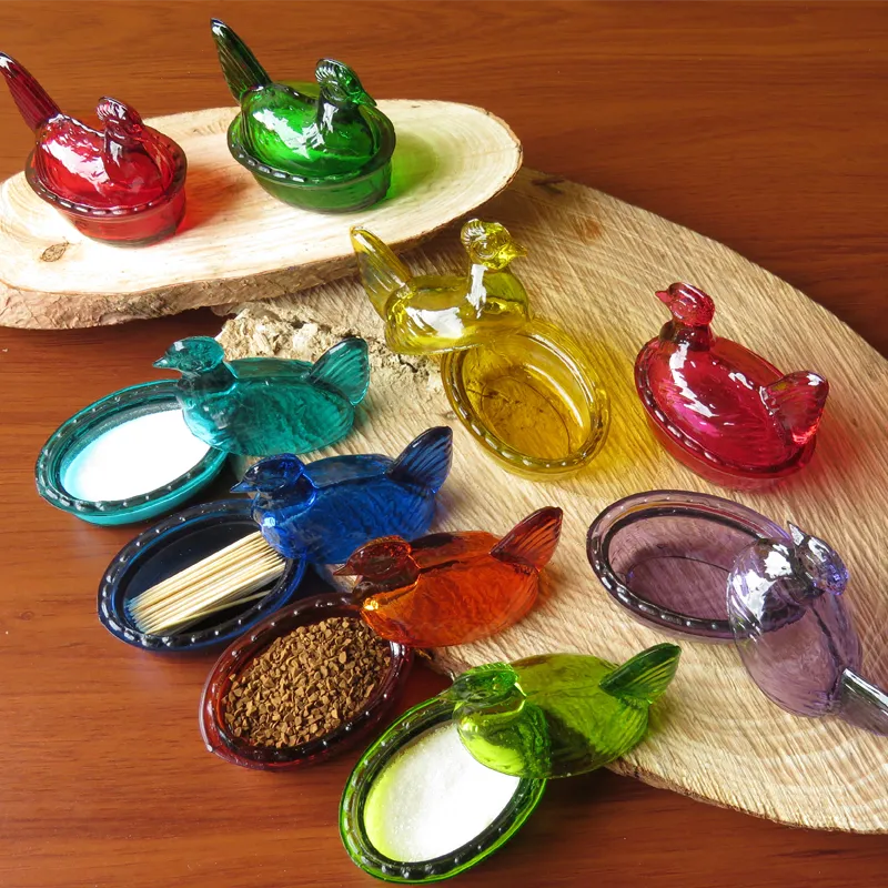 Colección de gallinas de vidrio artesanales de varios colores, mostrando sus múltiples usos como salero, palillero y recipiente para café sobre una mesa de madera.