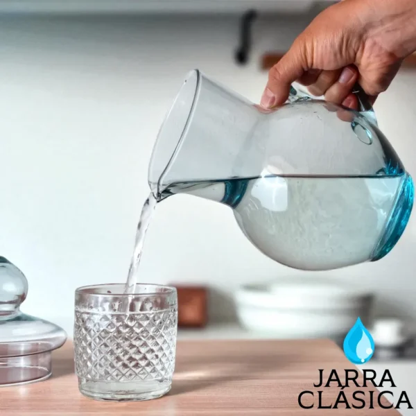 Mano sirviendo agua desde la Jarra Clásica de vidrio transparente en un vaso texturizado, en un ambiente de cocina.