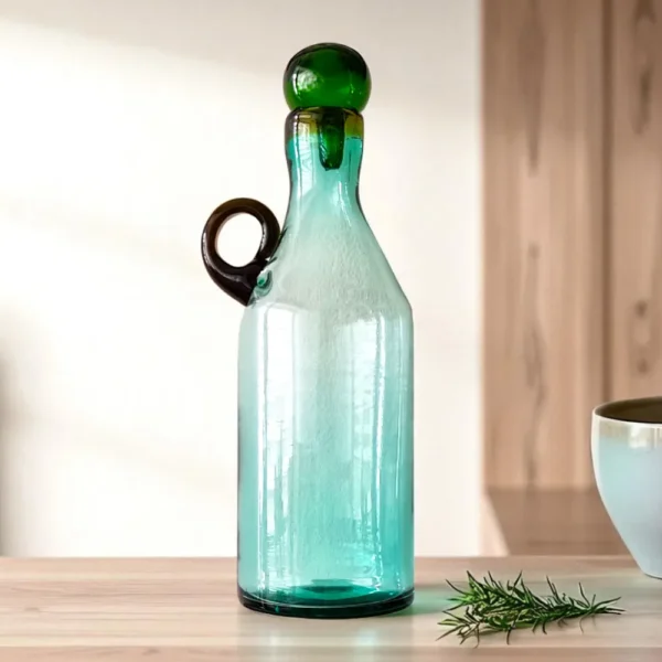 Botella de vidrio decorativa estilo mexicano, ambientada en una cocina sobre una mesa de madera con una rama de romero al lado.