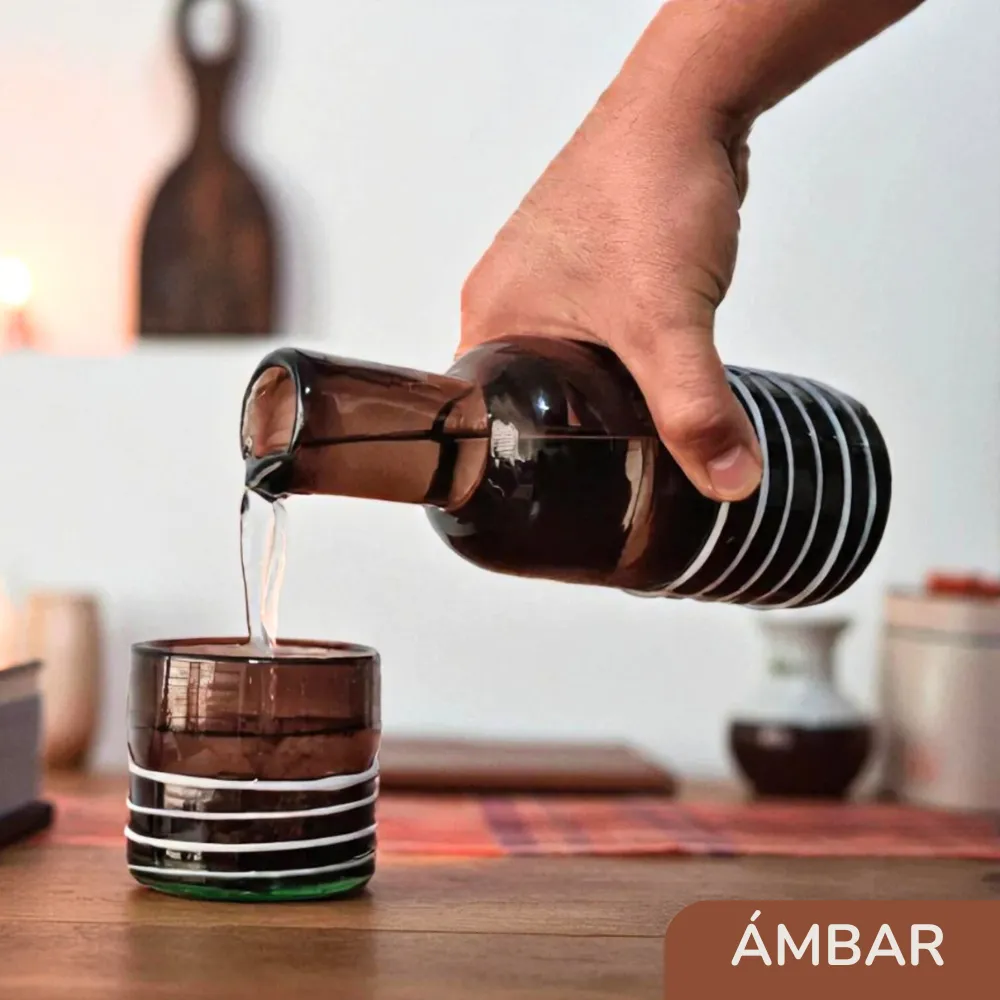 Mano sirviendo una bebida desde la botella de vidrio ámbar artesanal hacia su vaso sobre una mesa de comedor.