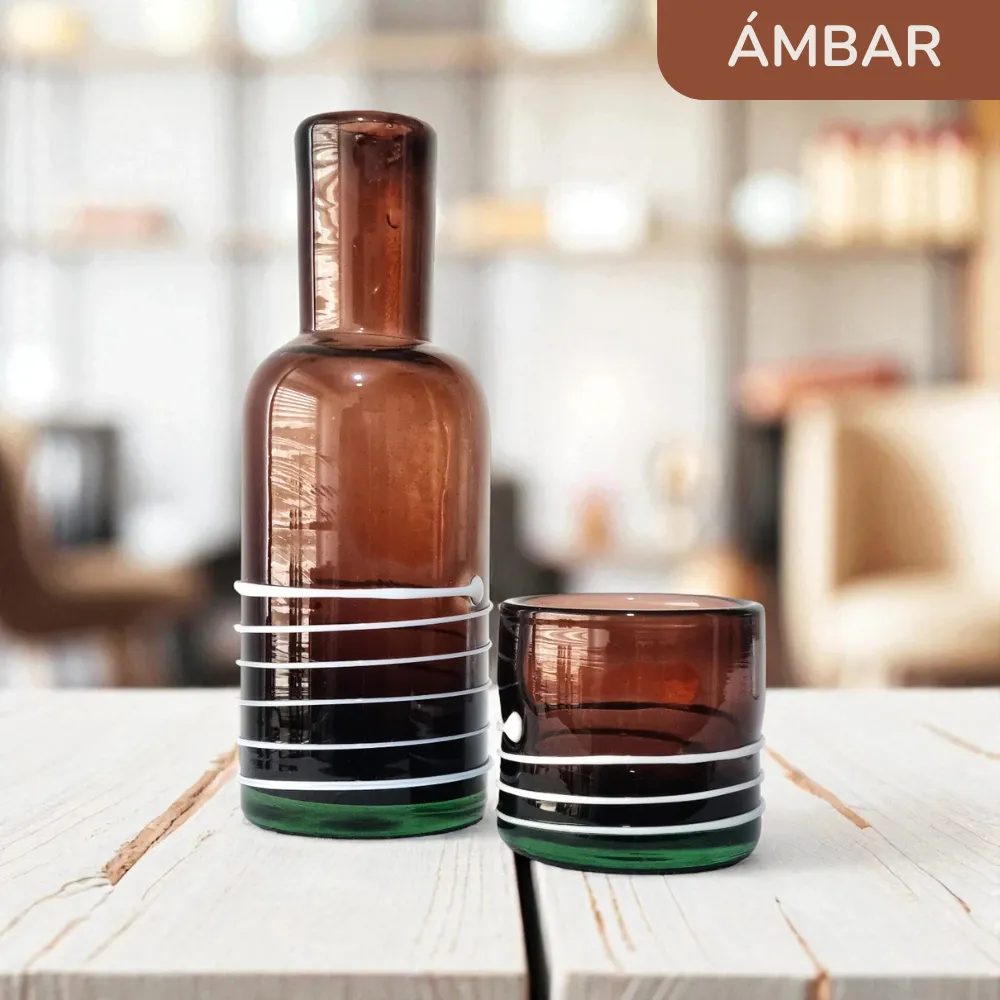 Set de botella y vaso artesanal de vidrio ámbar sobre una mesa de madera en un interior cálido y luminoso.