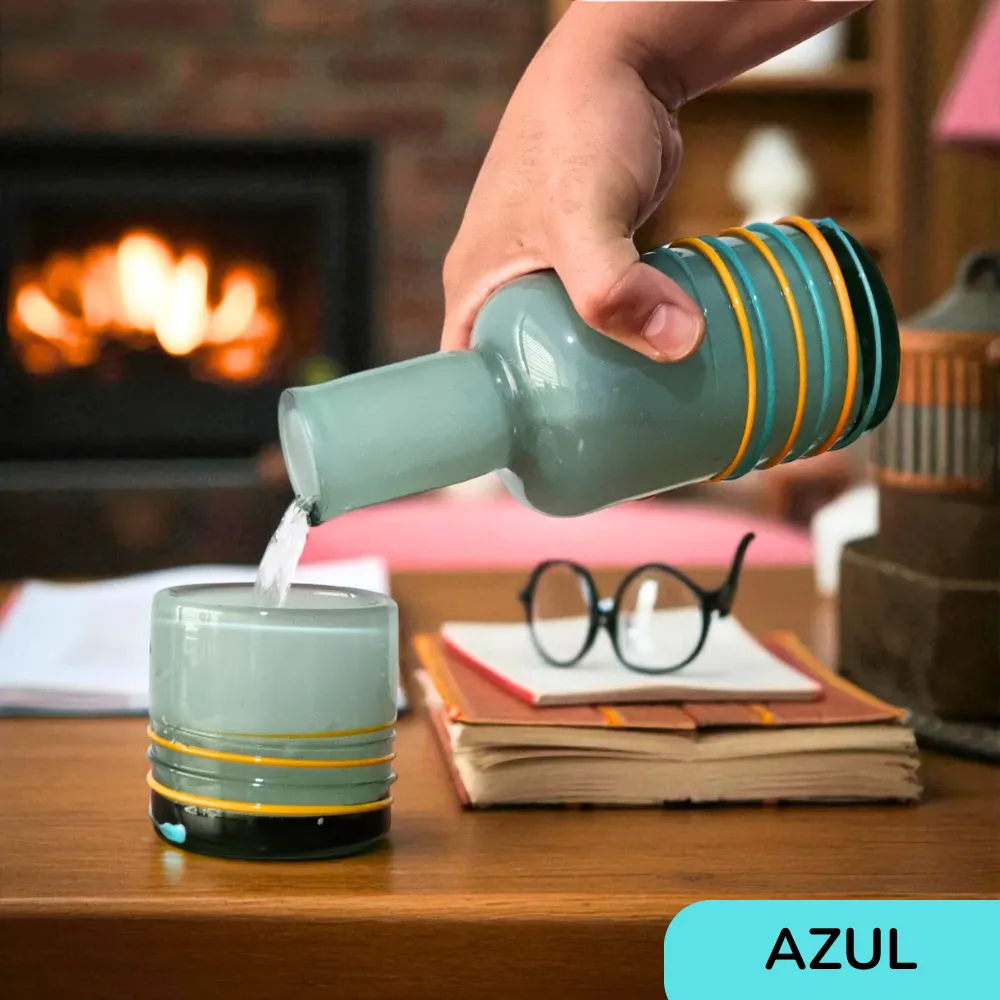 Mano sirviendo líquido desde la botella de vidrio artesanal azul en su vaso a juego, en un ambiente acogedor con una chimenea de fondo.