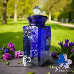 Dispensador de bebidas de vidrio azul cobalto, modelo México, en una mesa de jardín rodeado de flores moradas y rosadas.