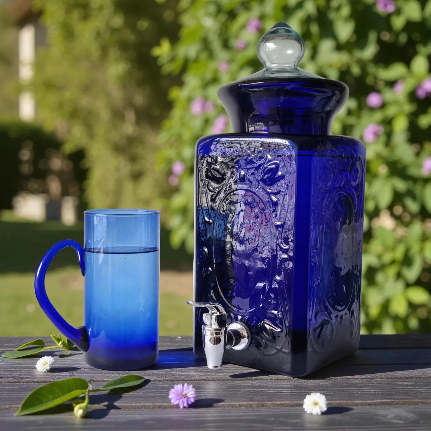 Home 34 Dispensador de bebidas de vidrio azul artesanal, modelo México, con su llave y un vaso azul a juego sobre una mesa de madera en un jardín.