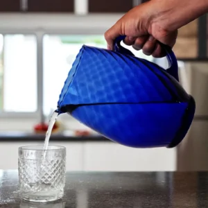 Mano sirviendo agua desde la jarra de vidrio estriada Chimy color azul cobalto en un vaso de vidrio sobre una encimera de cocina.