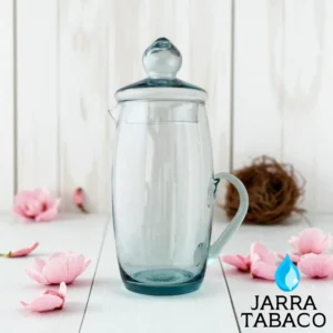 Jarra de vidrio Tabaco transparente con tapa, en una ambientación primaveral sobre una mesa blanca y rodeada de flores rosadas.